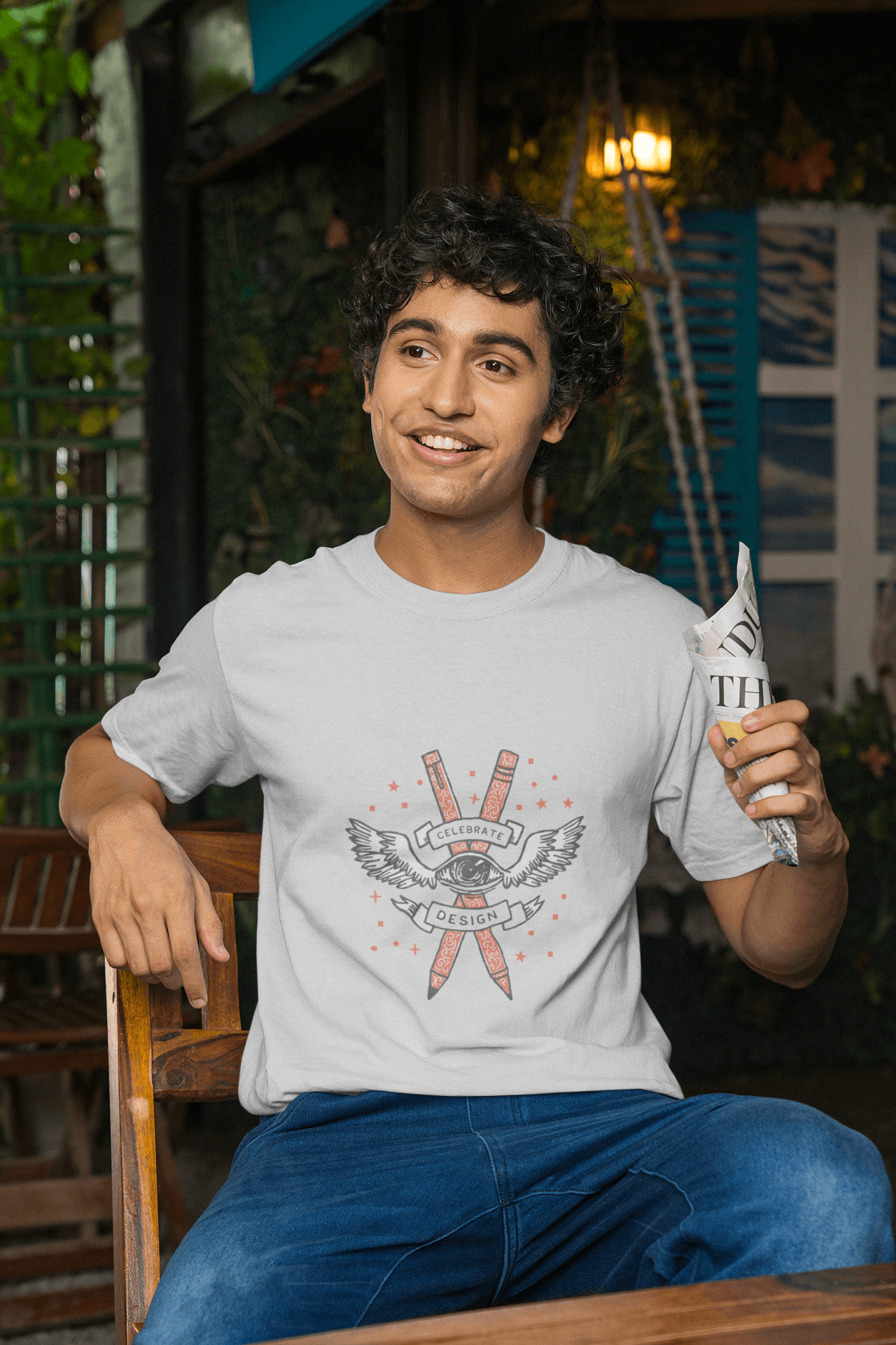 Man wearing Celebrate Design Unisex Tshirt with vibrant graphic print, sitting outdoors and smiling while holding a newspaper.