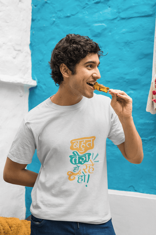 Person wearing a white unisex t-shirt with the text "BAHUT TEJ HO RAHE HO" eating a snack against a blue and white background