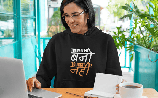 Woman wearing a Traveller Hooded Sweatshirt while working on a laptop in a vibrant café setting.