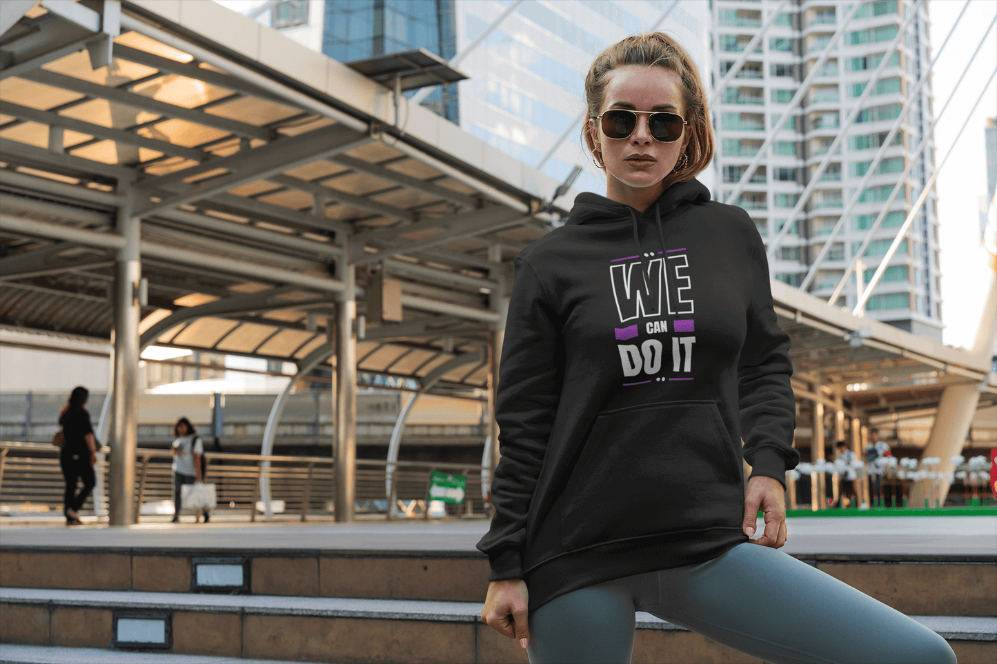 Stylish woman in black hoodie with "WE CAN DO IT" graphic, showcasing warmth and trendy fashion in an urban setting.