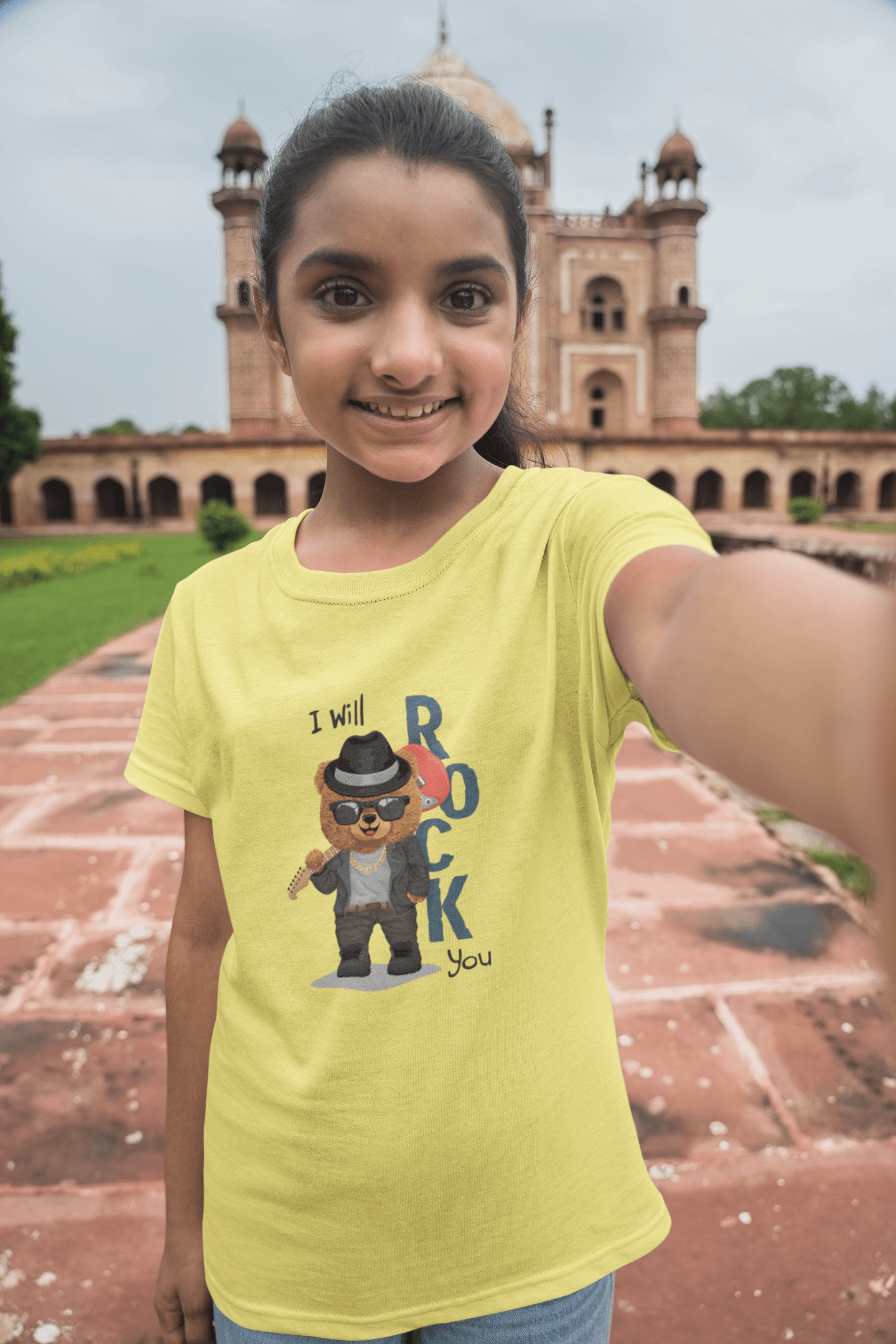 Girl wearing yellow "I Will Rock You" kids round neck t-shirt standing in front of a historic building