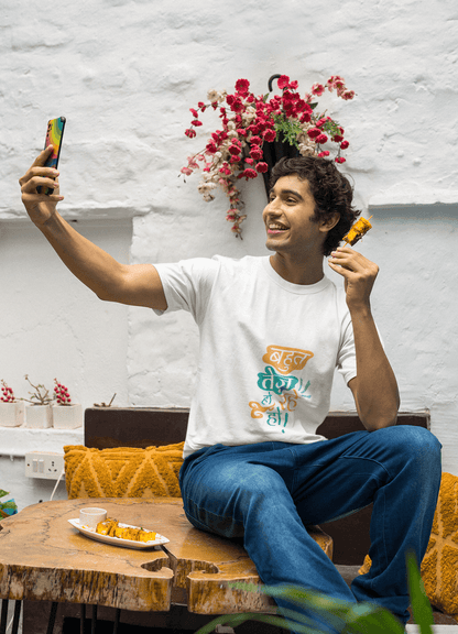 Man wearing BAHUT TEJ HO RAHE HO unisex t-shirt, sitting at a wooden table, taking a selfie while enjoying a snack, in a cozy indoor setting.