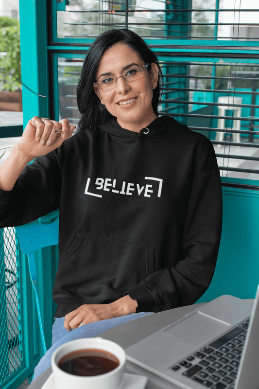 Woman in a black unisex hooded sweatshirt with "BELIEVE" design, sitting at a table with a laptop and coffee.