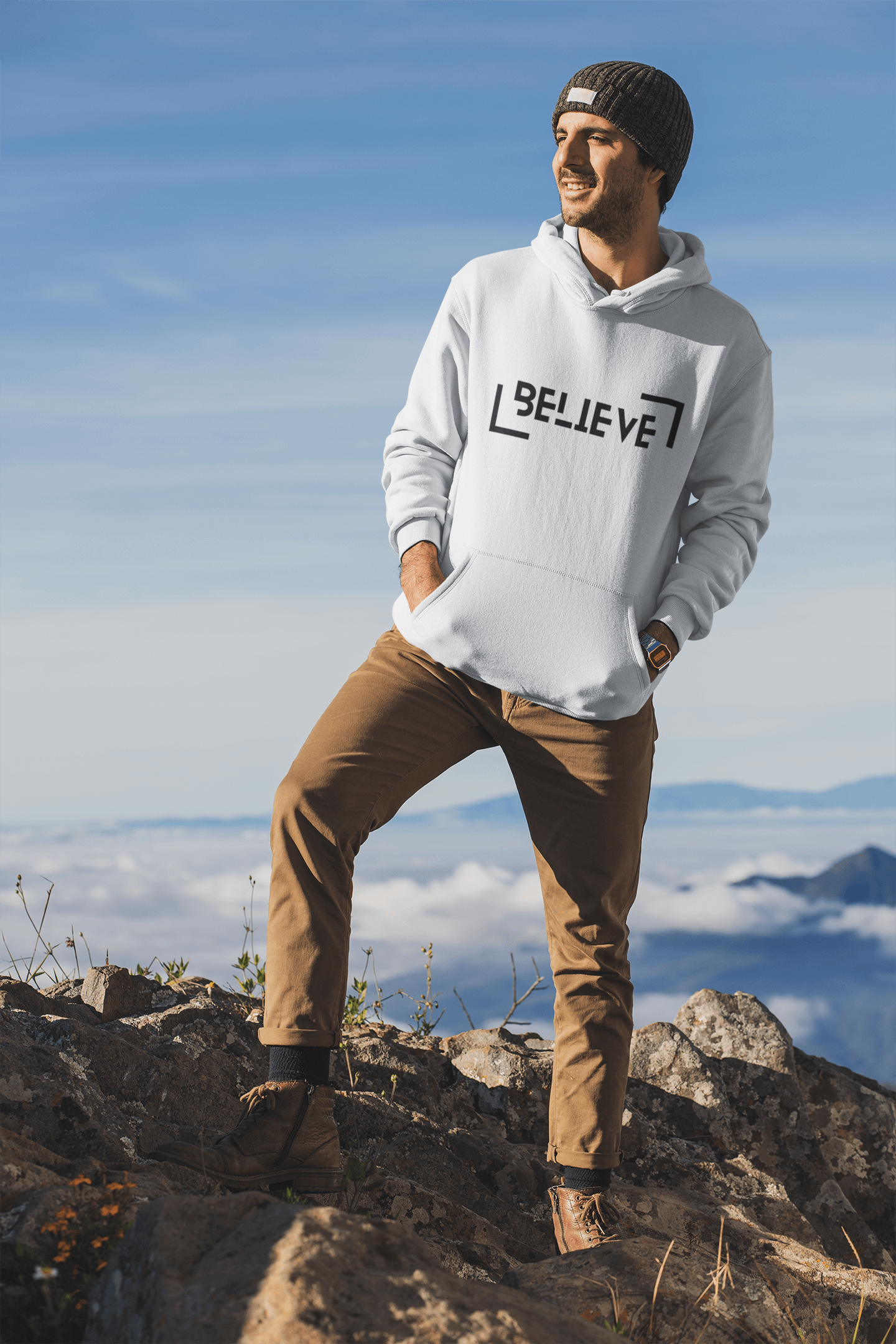 Man in a white Believe hooded sweatshirt standing on a rocky mountain top, showcasing a stylish winter look.