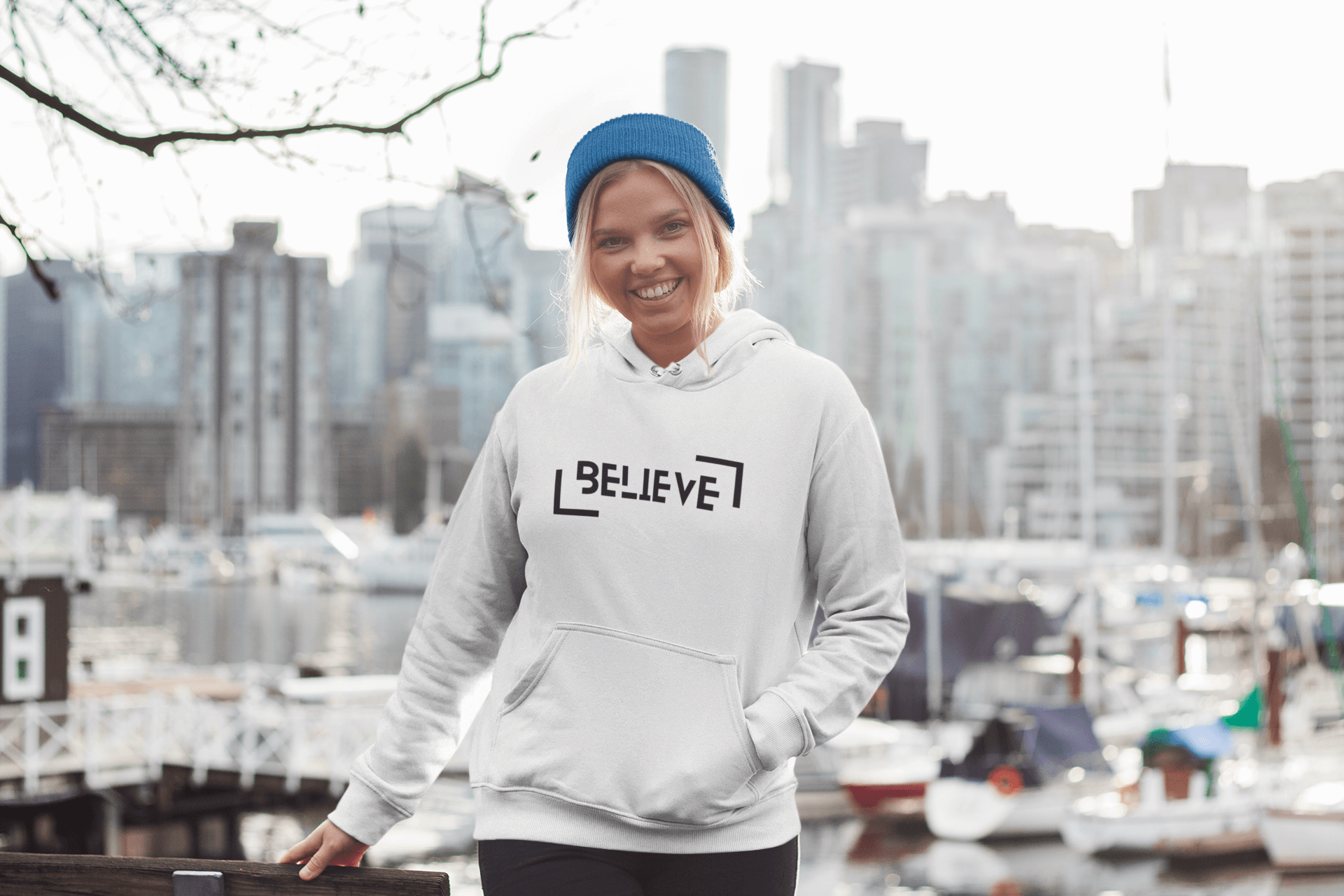 Woman wearing a stylish Believe hooded sweatshirt, standing by the waterfront with city skyline in the background.