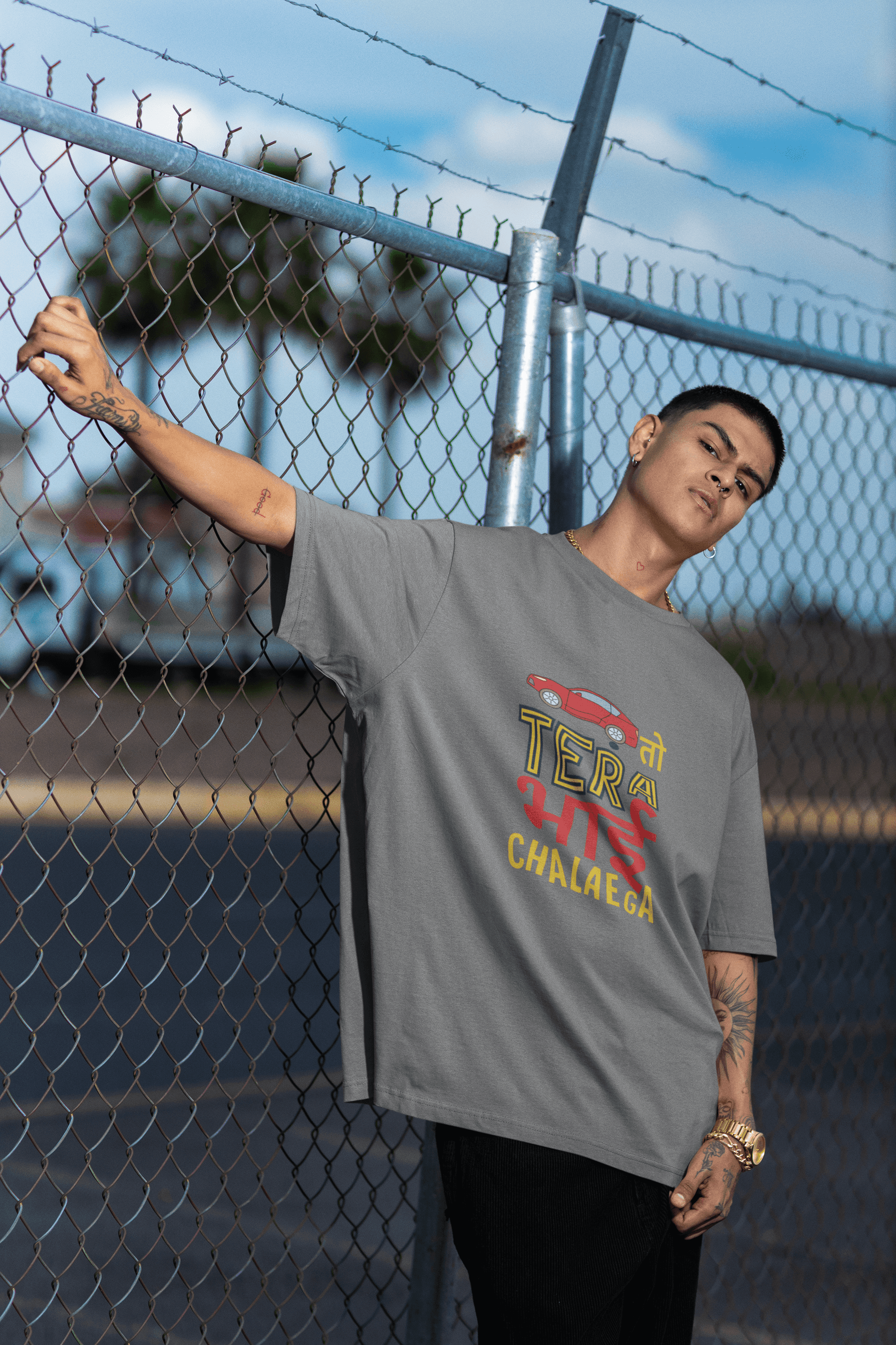 Person wearing a "GAADI TO TERA BHAI CHALAEGA" unisex T-shirt, gray shirt, against a chain-link fence background.